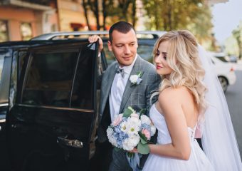 Beautiful and stylish newlyweds stand in the city on the background of the car. A stylish bridegroom, leaning against the door of the car, looks at a sweet bride with curly hair.